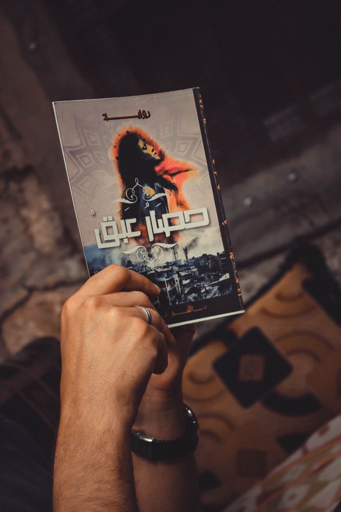 A person holds a book with an illustrated cover inside a cozy home in Syria.