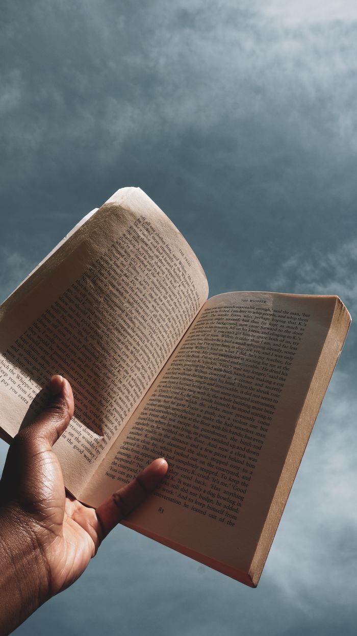 From below of crop anonymous person holding and reading opened textbook against cloudy blue sky on sunny day